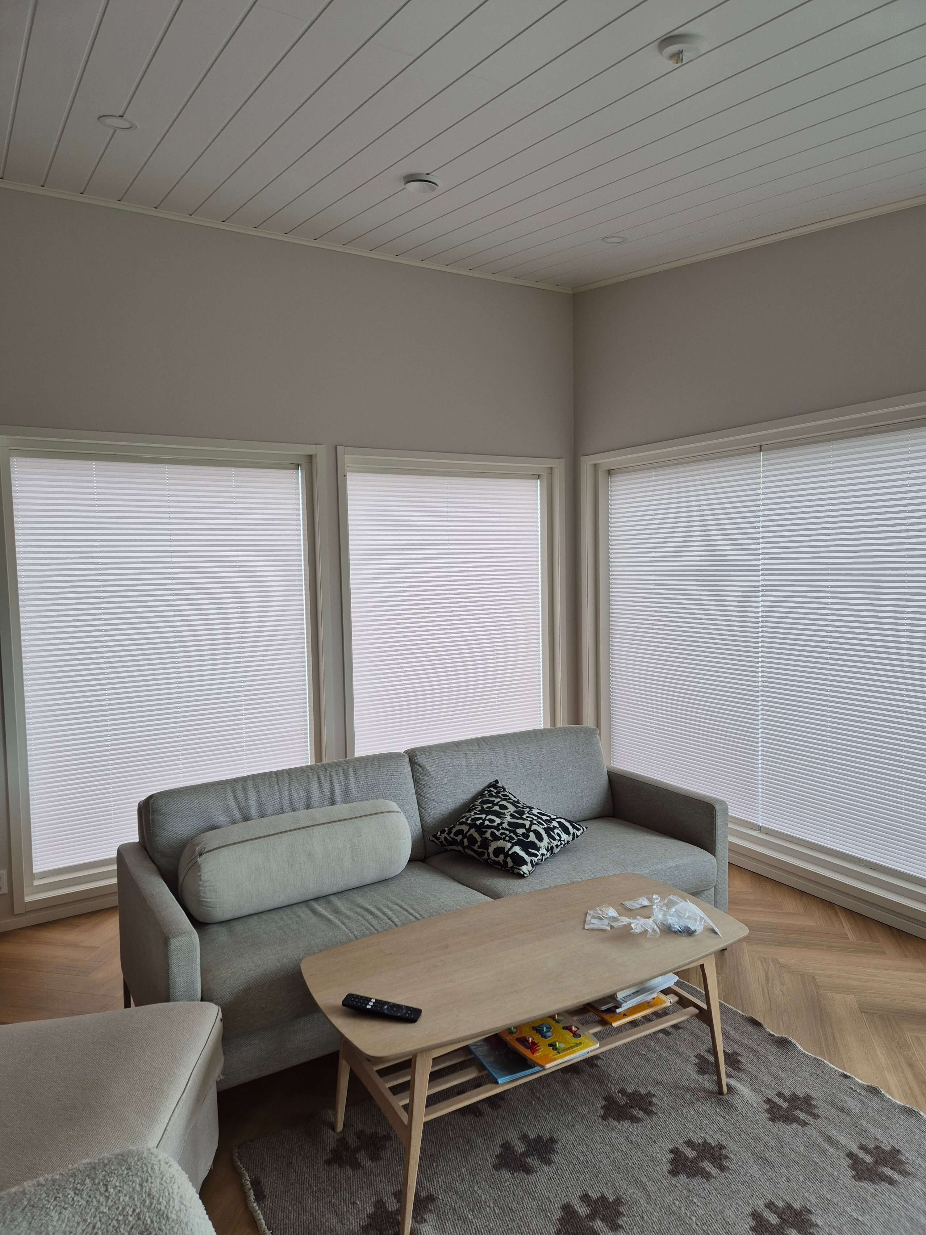 Moderni harmaa sohva, jossa kaksi koristetyynyä, puinen sohvapöytä ja ruskea matto valkoisten kaihtimien edessä.