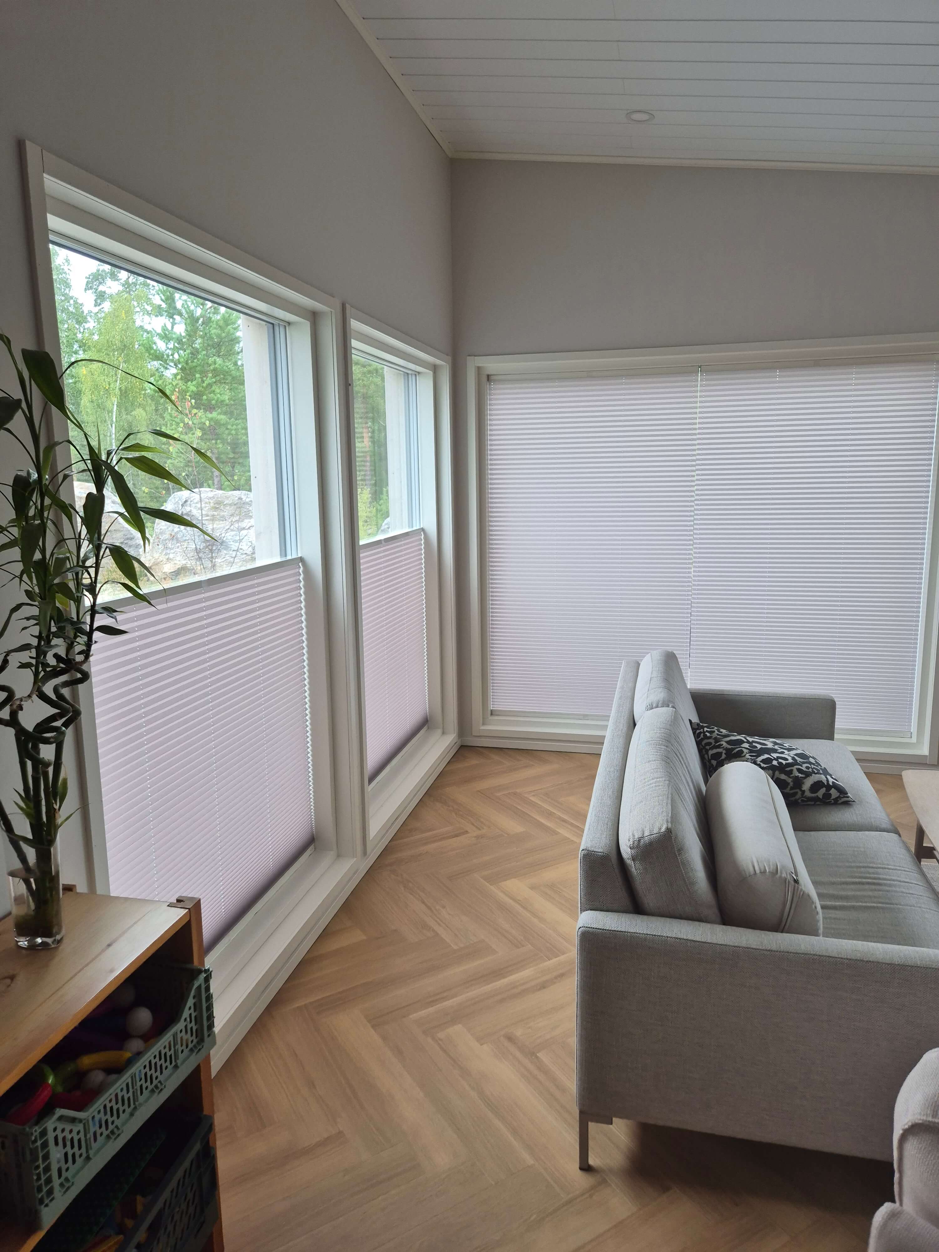 Valoisa olohuone, jossa on vaalea sohva, puulattia ja isot ikkunat, joissa puoliverhot.