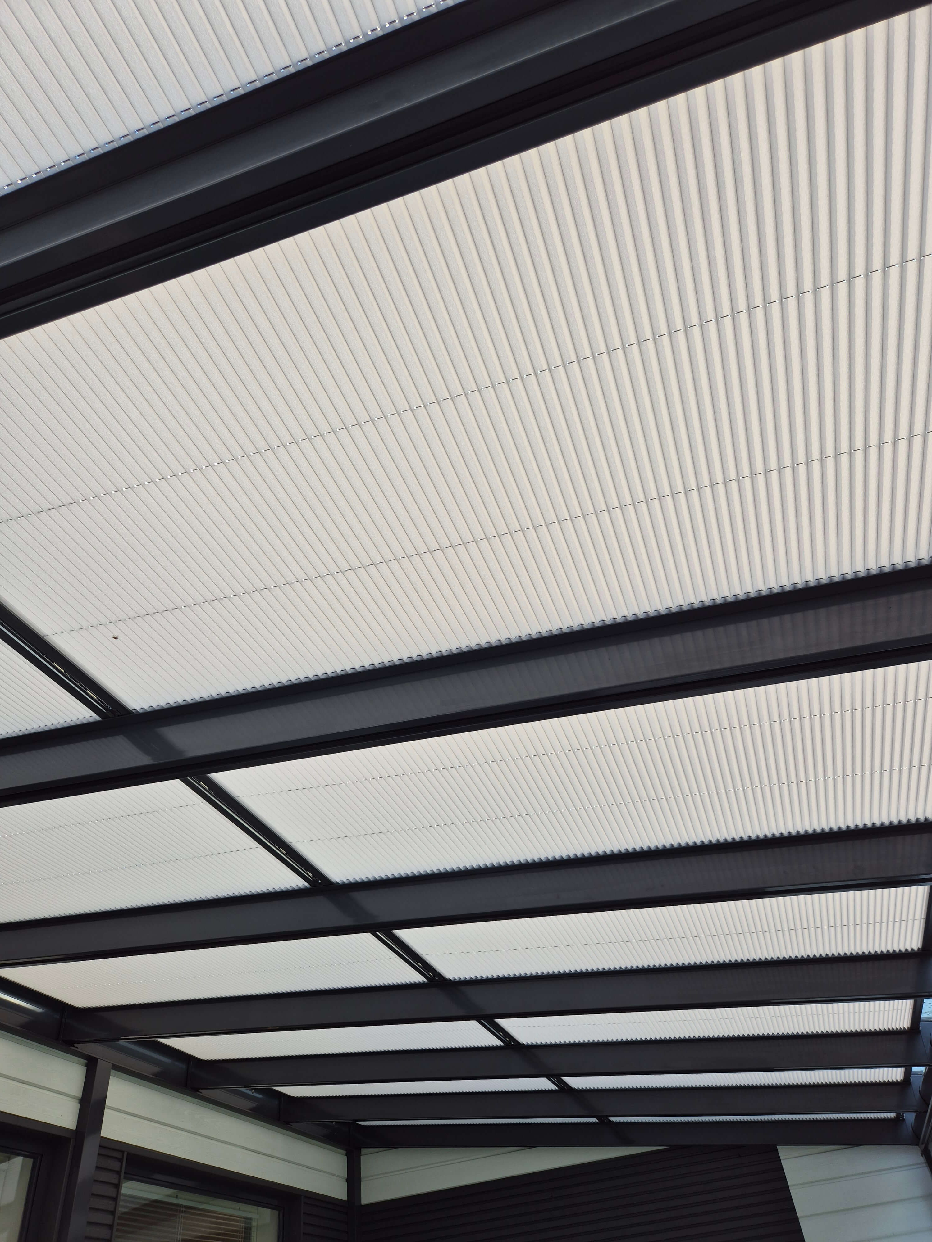 Rakenne, jossa on läpinäkyvä katto, jossa on mustat tukipalkit ja vaaleat vaakasuorat paneelit.