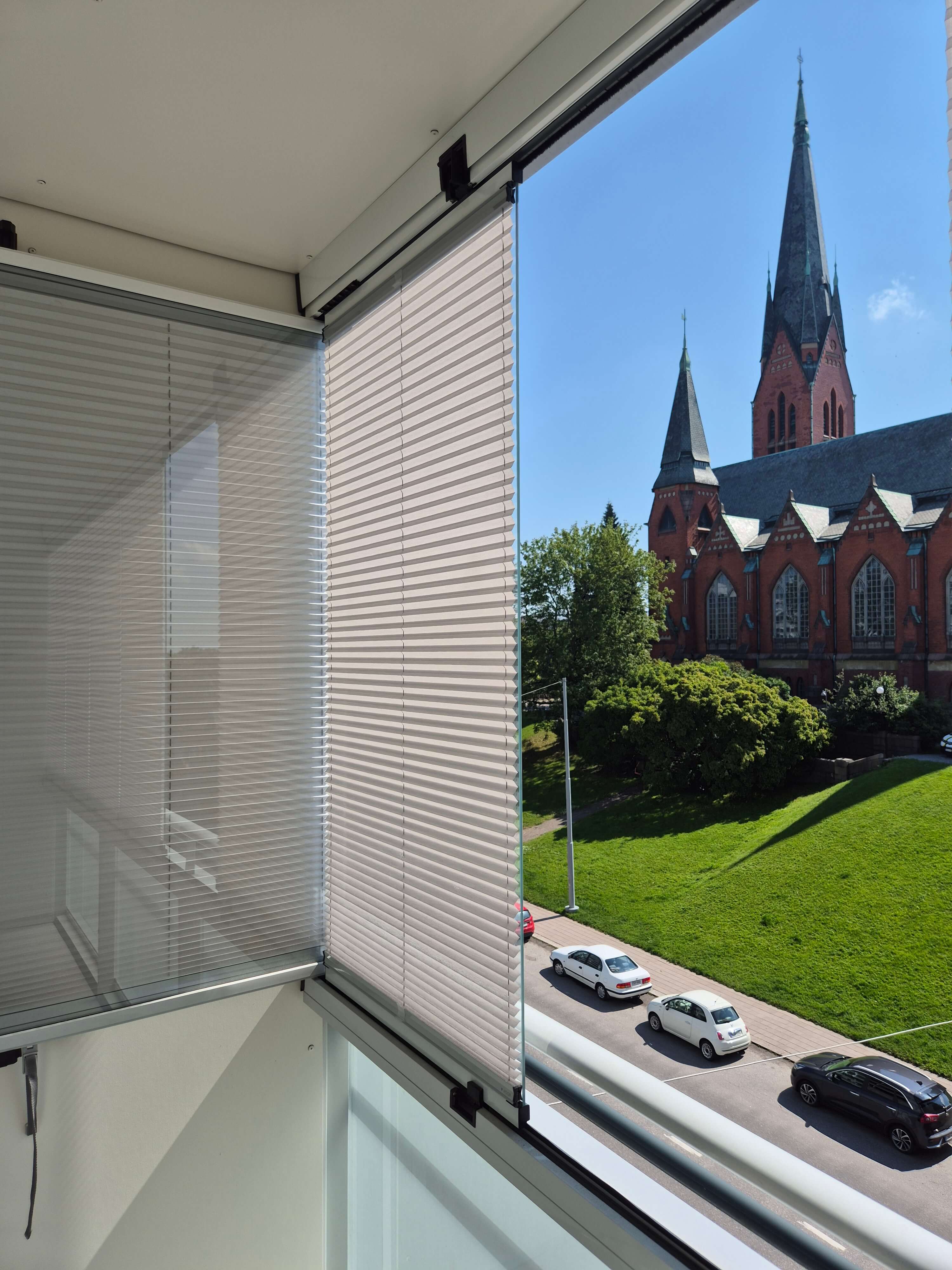 Näkymä parvekkeelta, jossa valkoiset sälekaihtimet ja taustalla punatiilinen kirkko sinistä taivasta vasten.