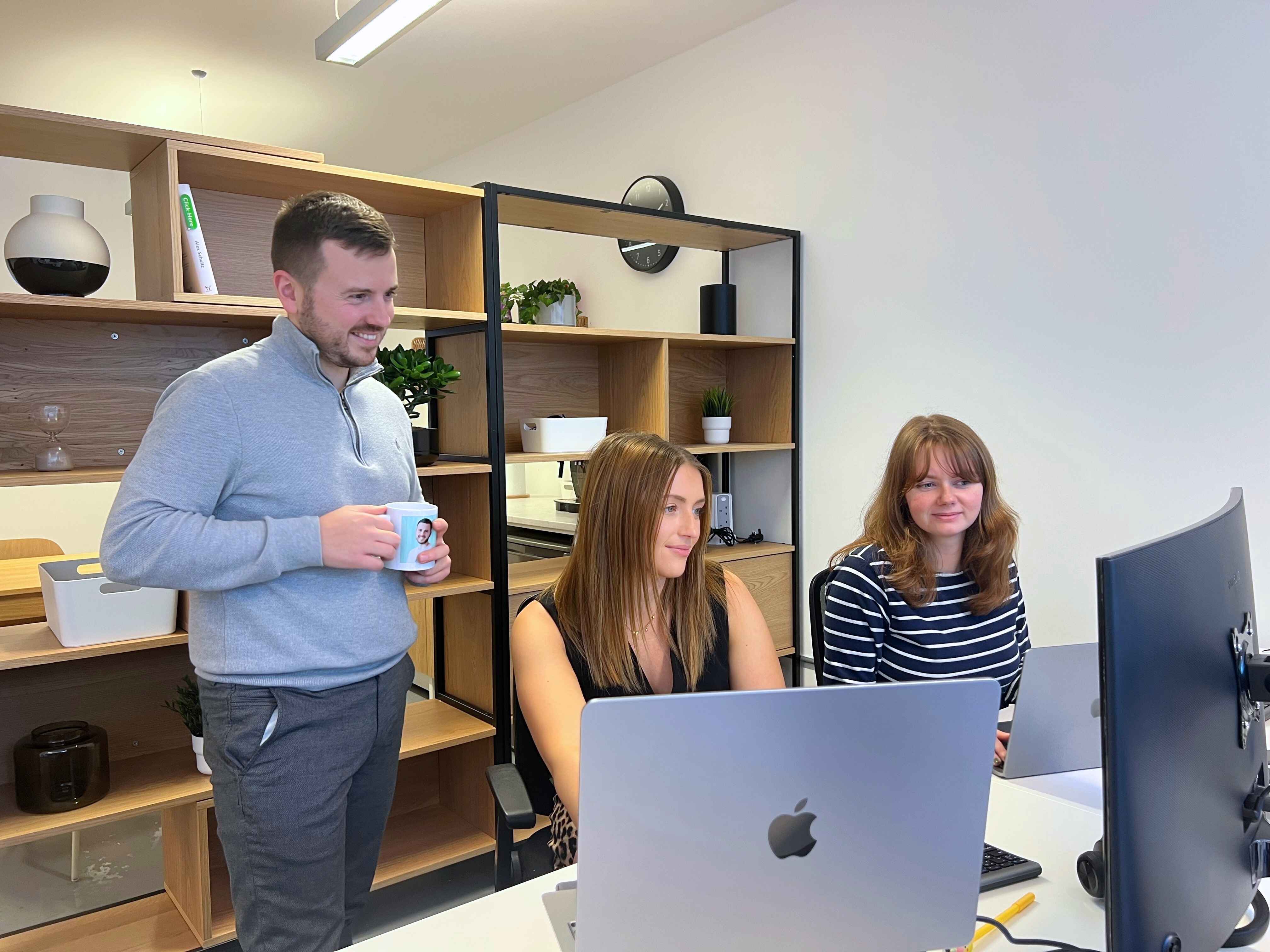 Two women sitting at a desk working on laptops while a man stands beside them holding a coffee mug in a modern office.
