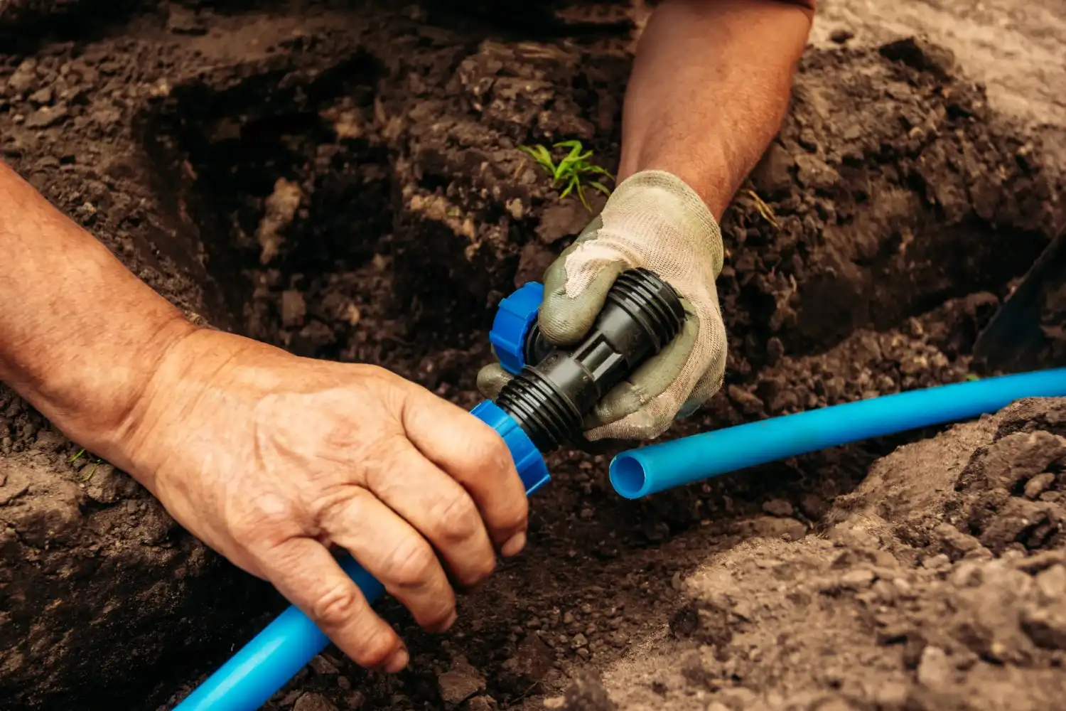 A person wearing a glove holds a black and blue compression fitting, preparing to connect a smaller blue water pipe in a dirt trench.