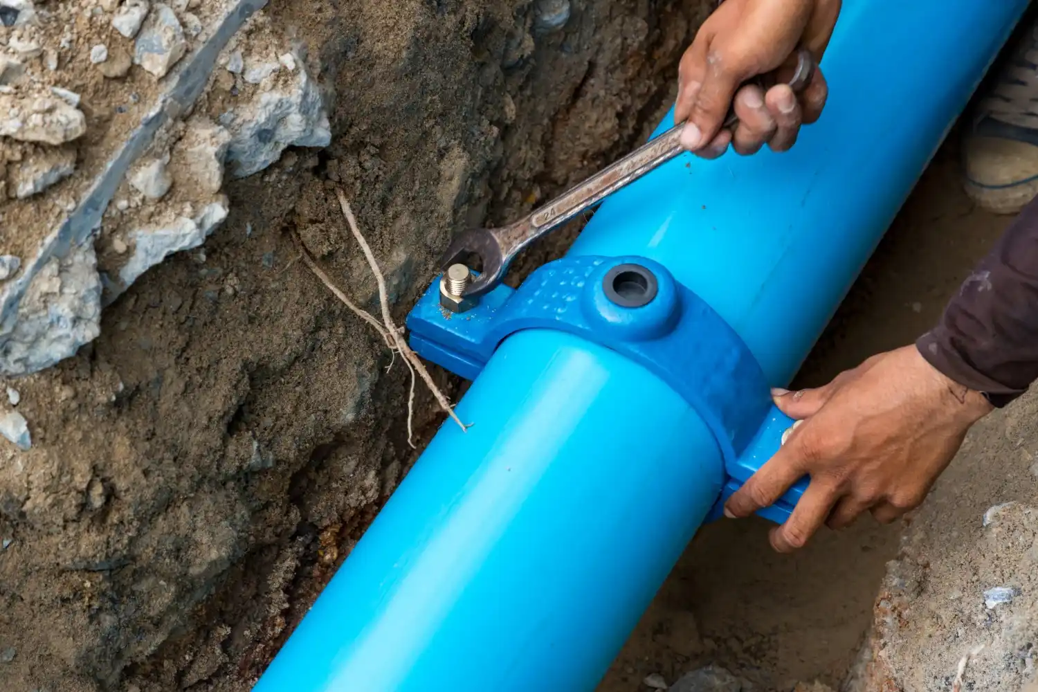 A close-up of a worker using a wrench to secure a blue saddle clamp onto a large blue pipe, with a second hand stabilizing the pipe.
