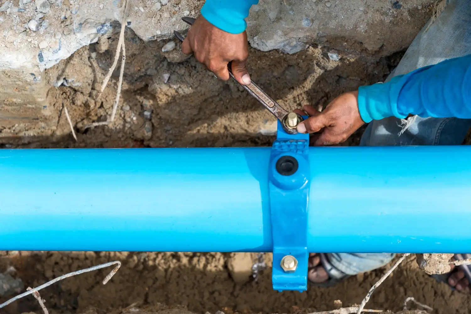 A person in a blue shirt uses a wrench to tighten a bolt on a blue clamp that's securing a large blue water pipe in an outdoor trench.