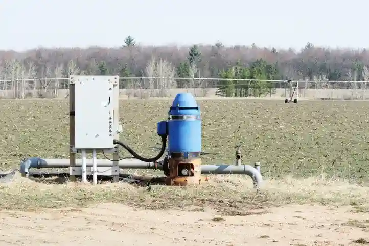 A large above-ground water pump with a blue casing and a rust-colored base sits in a dry, tilled field. The pump is connected to an electrical box and a network of gray pipes. In the distance, a large irrigation system on wheels stretches across the field. The background shows a line of bare trees under a hazy sky, suggesting a late fall or early spring setting.