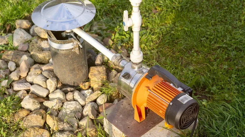 A small above-ground water pump with a bright orange electric motor and a silver body sits on a concrete slab. A white pipe with a valve extends from the pump toward a partially covered wellhead. The wellhead has a metallic, conical cover and is surrounded by a pile of large river rocks. The setup is located on a grassy lawn.