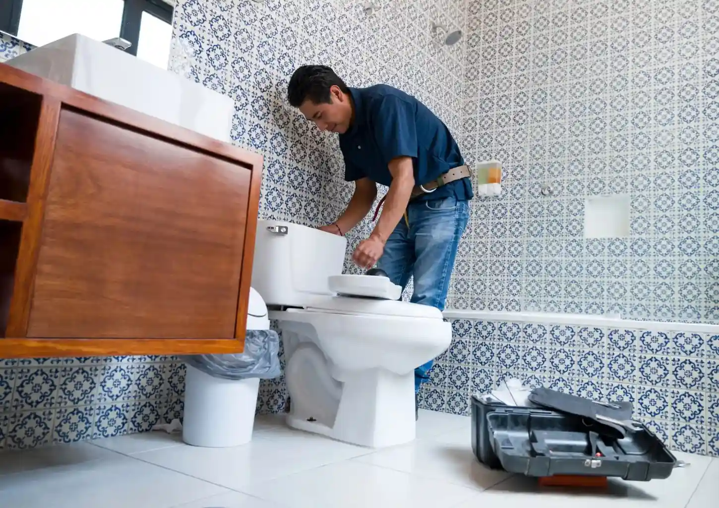 A man in a blue shirt and jeans repairs a toilet in a bathroom with blue and white patterned tile walls.