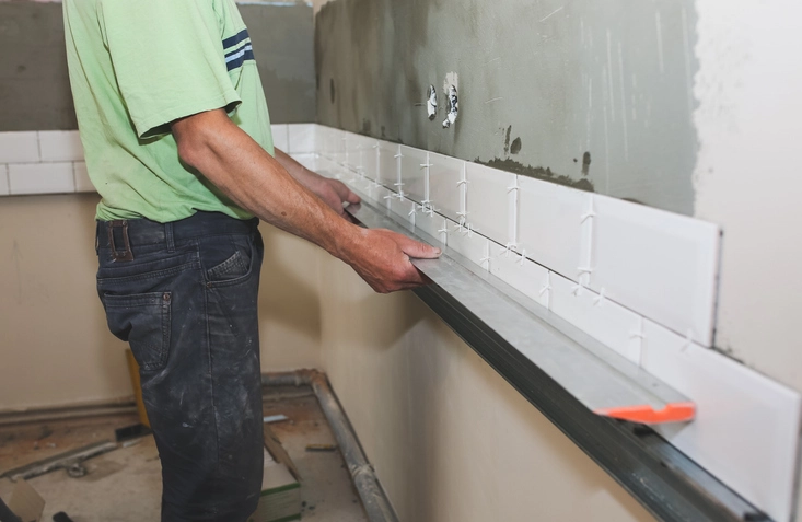 A person wearing a light green shirt and dark jeans is working on a wall, holding a long, silver metal leveling tool. They are installing white, rectangular subway tiles on the lower part of the wall, which appears to be a backsplash or a feature wall in a kitchen or bathroom. The tiles are held in place with spacers, and the rest of the wall is unpainted and unfinished.