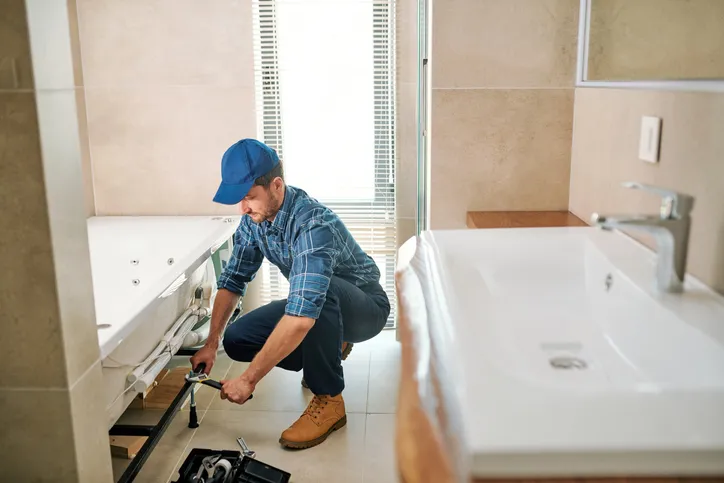 A plumber in a blue plaid shirt and cap kneels by a white bathtub, using a wrench to work on pipes.