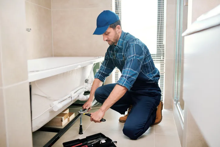 A plumber in a blue plaid shirt and cap kneels, using a wrench to work on pipes under a bathtub.