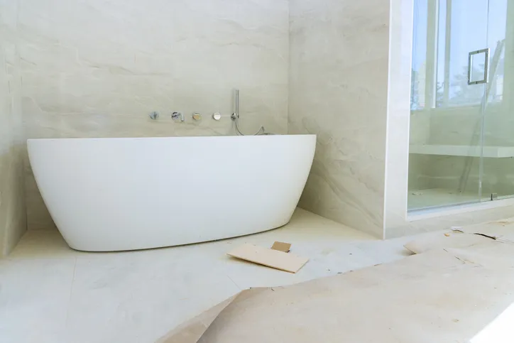A modern white freestanding bathtub is placed in a spacious, tiled bathroom with a large window and a glass shower stall.