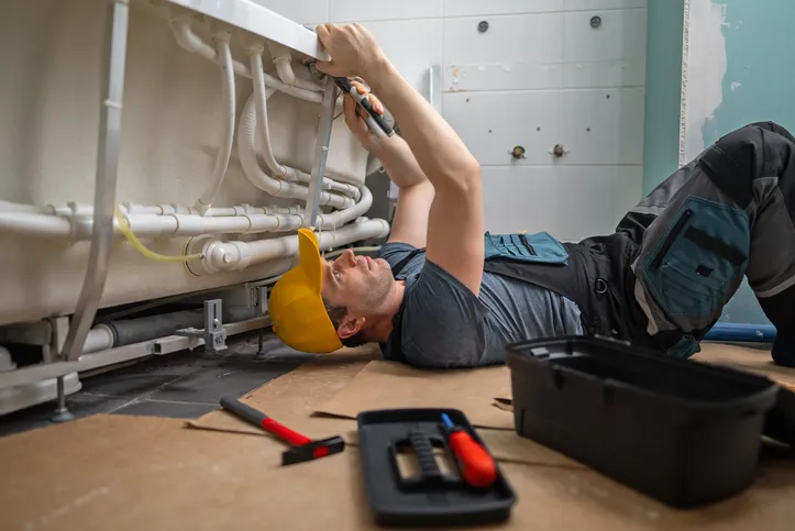 A plumber lies on their back on the floor, working on pipes under a bathtub with a wrench. A yellow hard hat is on the floor nearby.