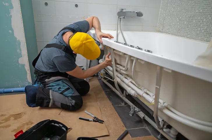 A plumber in a blue shirt, overalls, and a yellow cap kneels on a brown cloth, working on the pipes and wiring underneath a white bathtub.