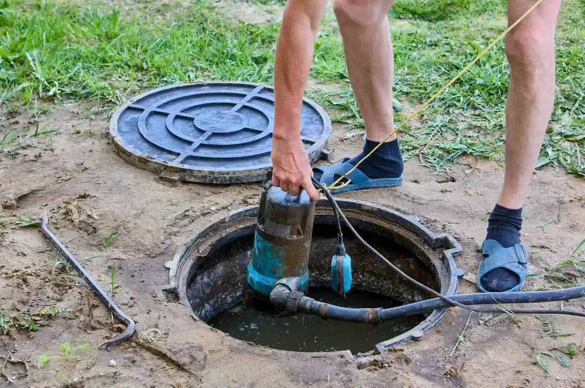 A person wearing sandals and socks is shown from the waist down, pulling a small, dirty pump out of an outdoor sewage or septic pit.