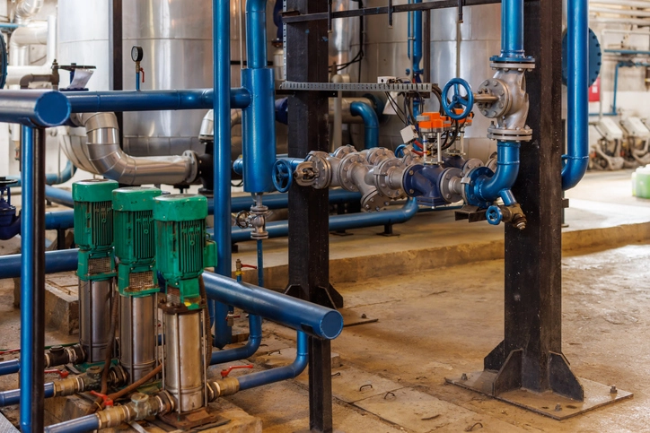 A wide-angle, industrial shot of a large-scale pumping system. The foreground features a trio of green, vertical booster pumps connected by a network of blue and silver pipes with numerous valves and gauges. The entire system is supported by heavy-duty black metal beams. The background is a mix of other pipes, tanks, and industrial equipment in a factory or plant setting.