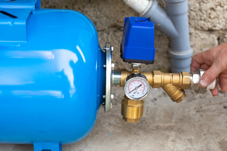 A close-up of a blue residential pressure tank connected to a water system. A hand is visible on the right, adjusting a brass pipe fitting. A pressure gauge with a white face and a blue pressure switch is mounted on top of the brass fittings, which connect to the tank. The tank and pipes are situated in a utility space, with a concrete floor and a rough, textured wall in the background.
