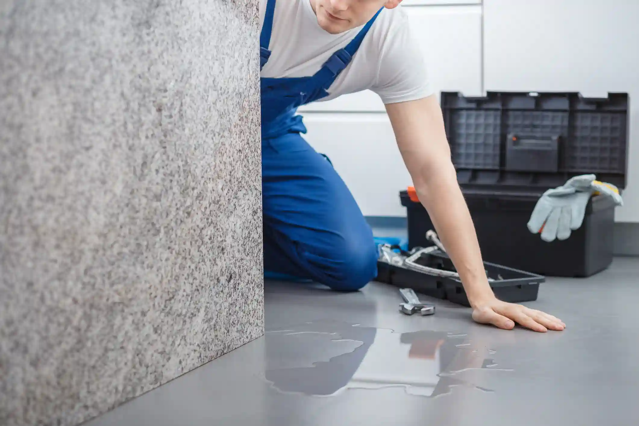 A young plumber, wearing a white shirt and blue overalls, is on his knees, reaching into a tight space behind a tiled kitchen island or counter. A puddle of water is visible on the grey floor, suggesting a leak. A black toolbox with a pair of gloves and a wrench is on the floor beside him.