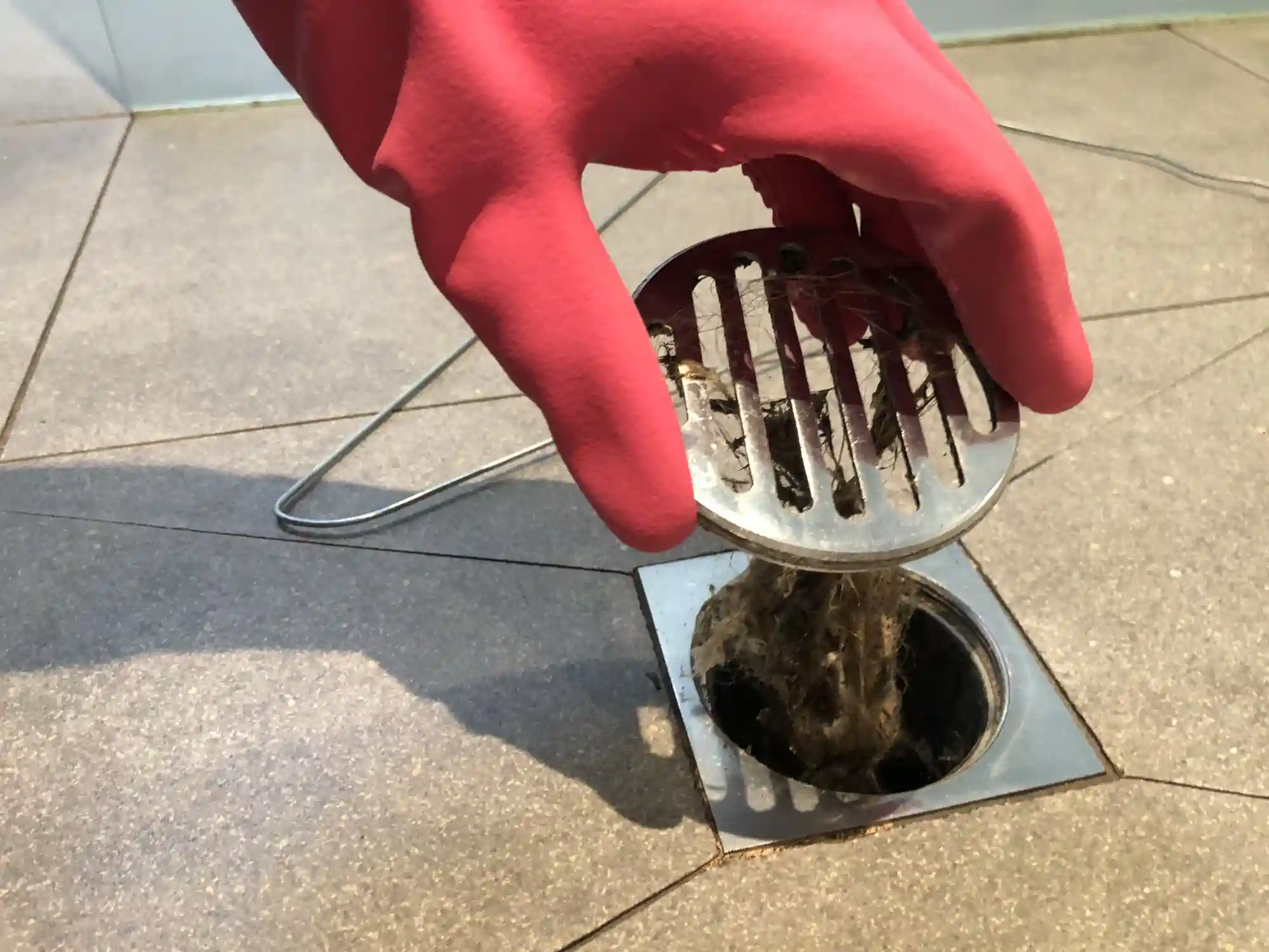 A close-up shot of a gloved hand in a bright red glove, holding up a round, metal drain cover. A large, repulsive clump of hair and grime is pulled from a floor drain, highlighting a common cause of clogs. The floor is tiled with light-colored, square tiles.