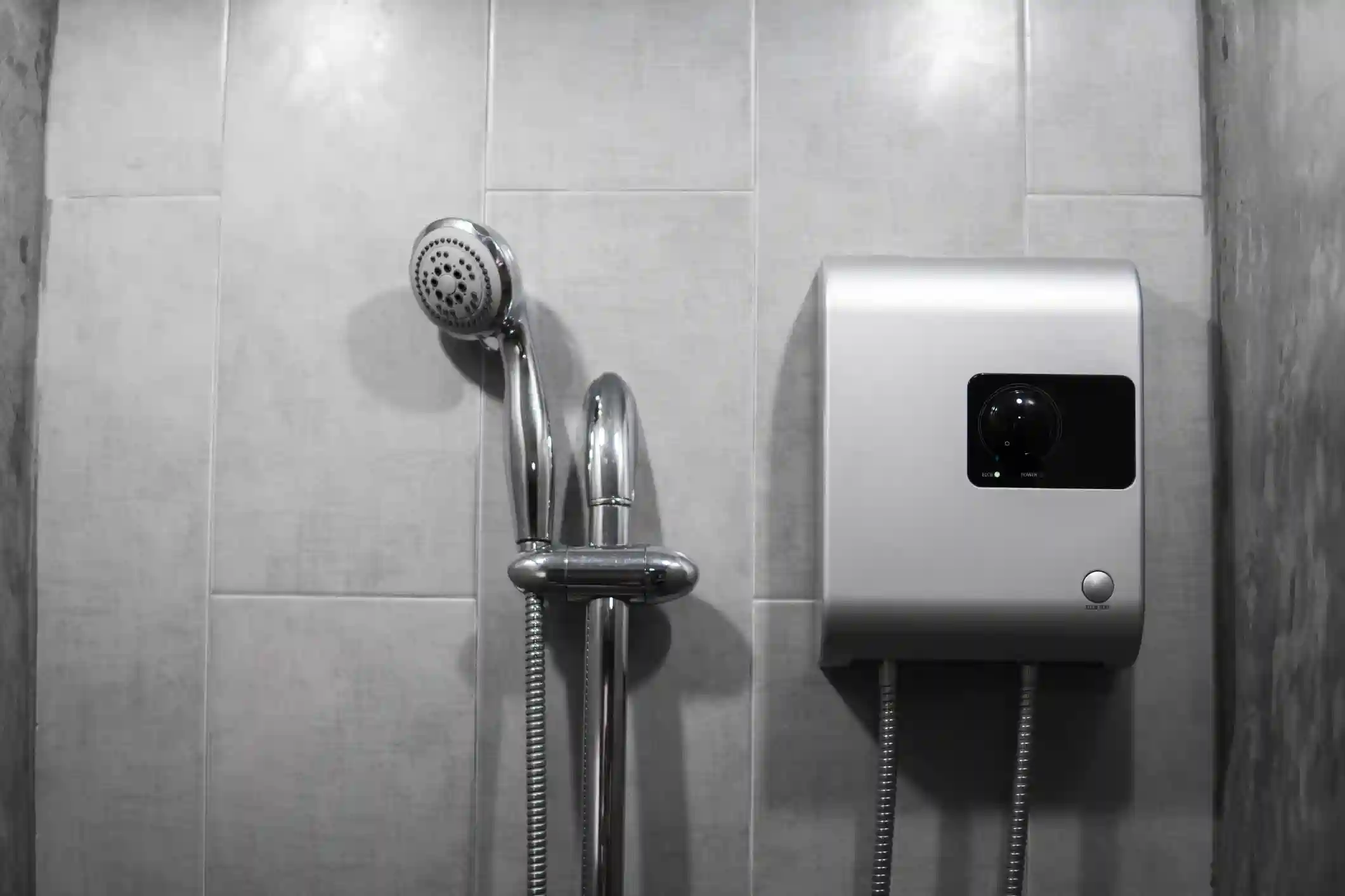 a compact, tankless water heater mounted on a tiled wall in a shower. The unit is silver and square-shaped with a black control panel and a single button at the bottom. A flexible metal hose connects to the bottom of the unit. A chrome shower head with a flexible hose and a mounting bracket is visible to the left. The wall is a mix of light gray tiles and concrete.