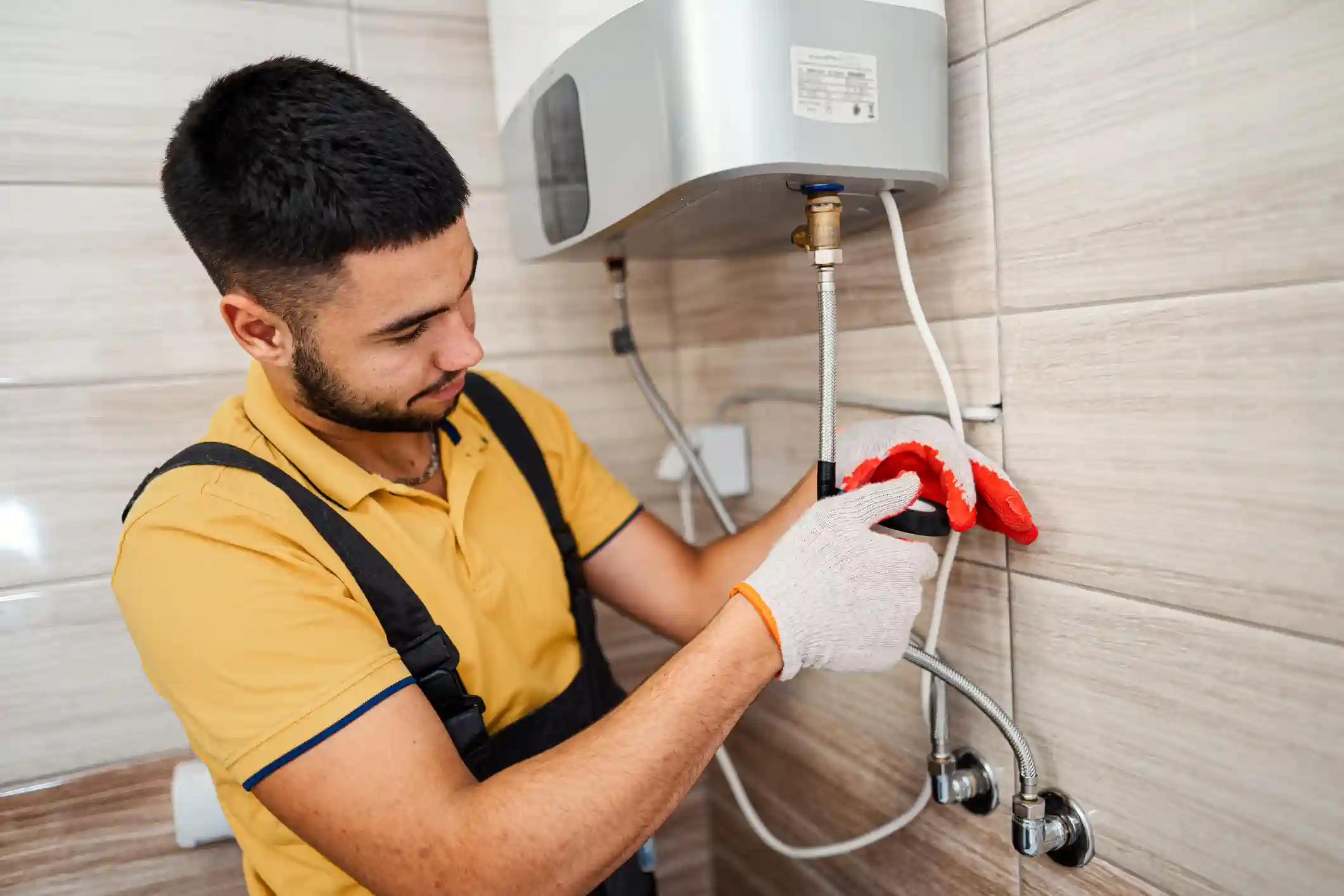 A male technician with a beard is installing a tankless water heater. He is wearing a yellow shirt, black overalls, and a pair of white gloves with red accents. He is holding and connecting a flexible metal water line to the bottom of the silver and white water heater unit. The unit is mounted on a light gray tiled wall.