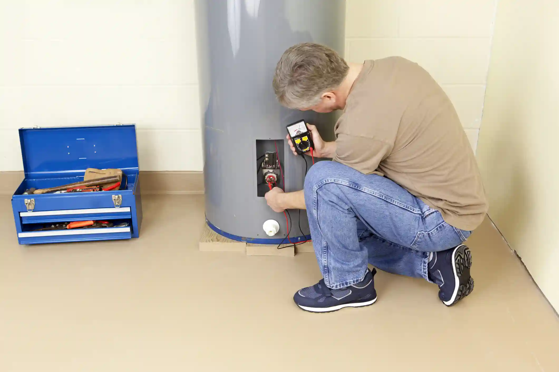 A repairman is crouched down, checking the electrical components of a large, gray tank water heater. He is using a multimeter with red and black probes to test the wiring inside an access panel on the side of the tank. A blue metal toolbox with various tools is visible on the floor to his left. The room has a light-colored floor and a white-walled background.