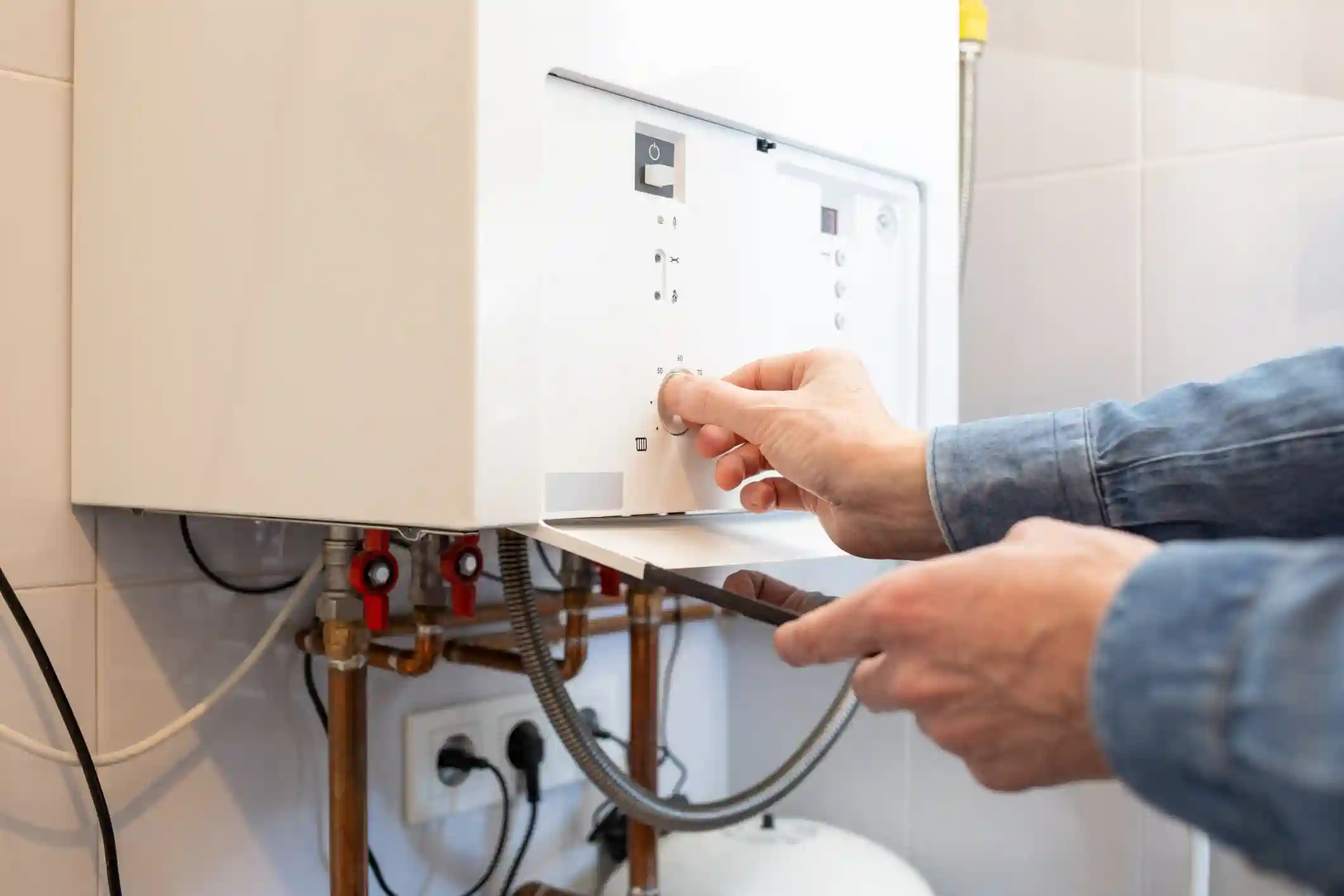 A person is adjusting the temperature dial on a white tankless water heater. The unit is mounted on a tiled wall, and its front panel is open, revealing a series of knobs and a control board. Copper and other pipes, as well as electrical outlets, are visible below the unit. The person is using one hand to turn a knob and the other to hold a tool.