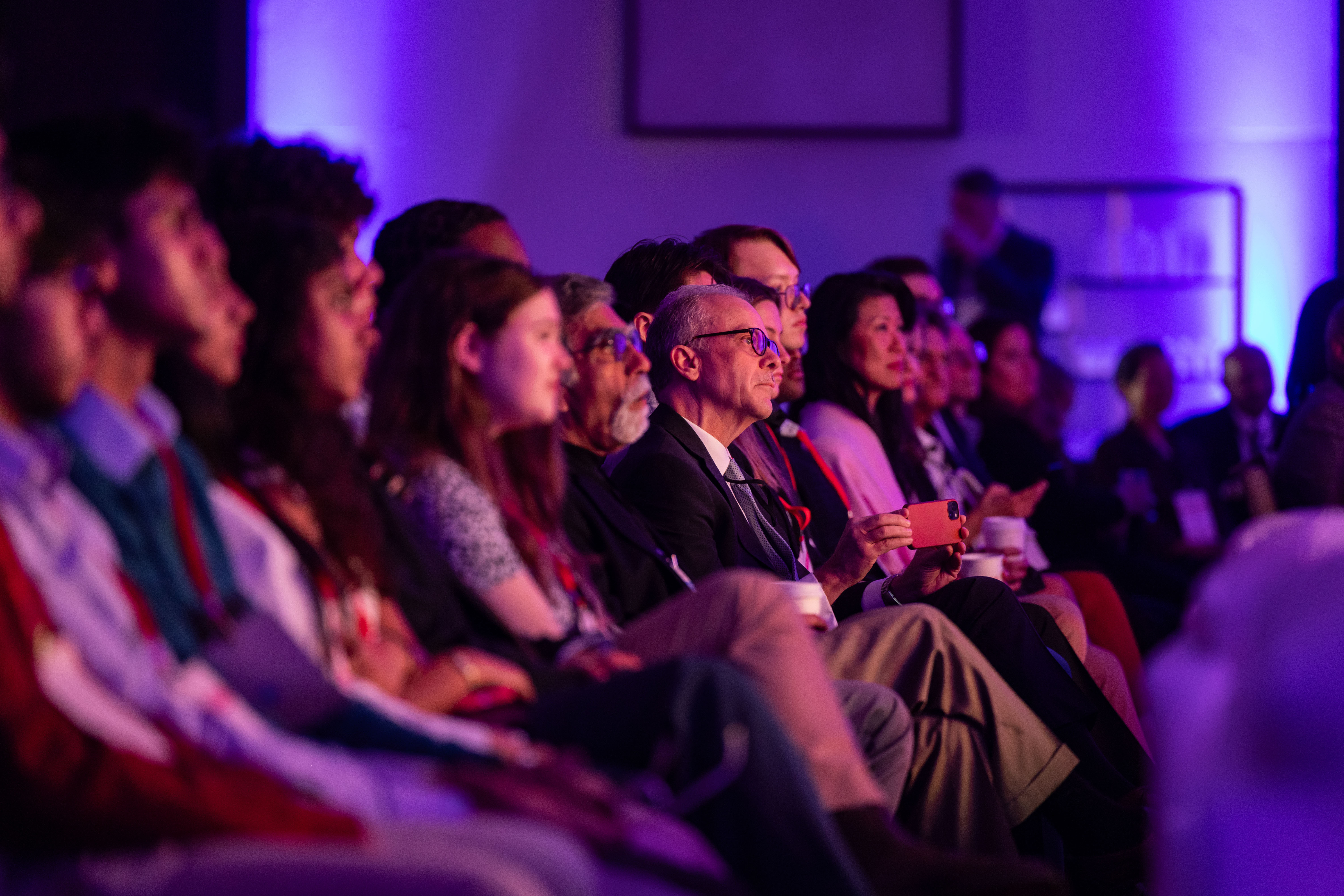 A diverse group of people seated closely in rows at an indoor event, attentively watching a presentation or speaker, illuminated by purple-blue lighting. One man in the center holds a smartphone in his hand while others hold cups, all facing towards the unseen stage.