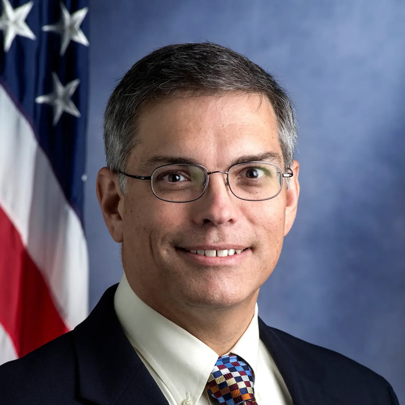 Smiling man with short salt-and-pepper hair and glasses wearing a dark suit, light shirt, and colorful patterned tie, with an American flag and blue background behind him.