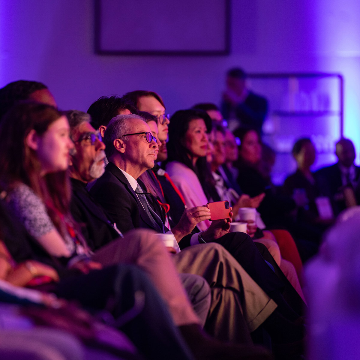 Audience members seated in rows attentively watching an event, with a man in the center holding a smartphone and others holding coffee cups in a room lit with purple lighting.