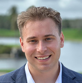Smiling young man with short curly hair wearing a blue blazer and white shirt standing outdoors with water and greenery in the background.