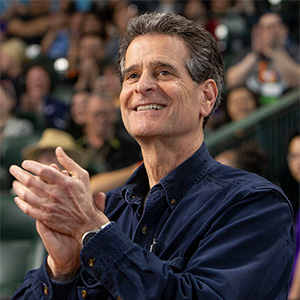 Man with dark hair and a navy shirt smiling and clapping hands in an indoor setting with blurred people in the background.