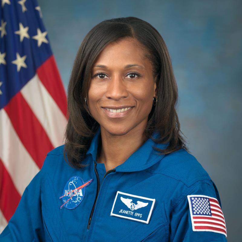 Portrait of a woman in a blue NASA flight suit with patches, including one with her name, Jeanette Epps, and an American flag patch on her shoulder, standing in front of a blurred American flag.