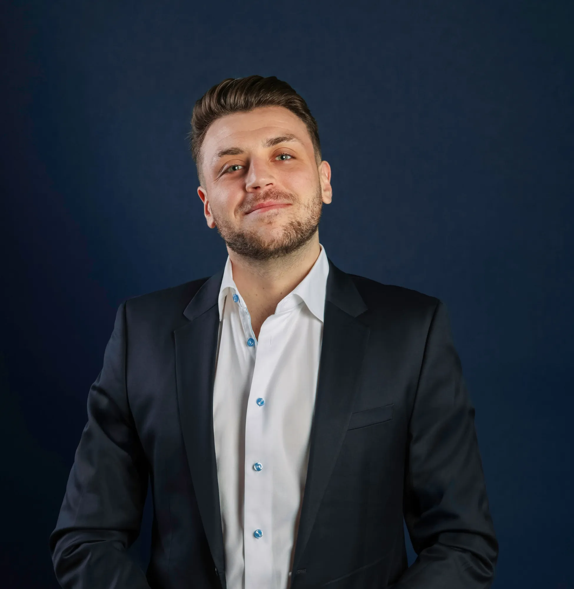 Smiling man with short hair and beard, wearing a black suit jacket and white shirt with blue buttons, standing against a dark blue background.