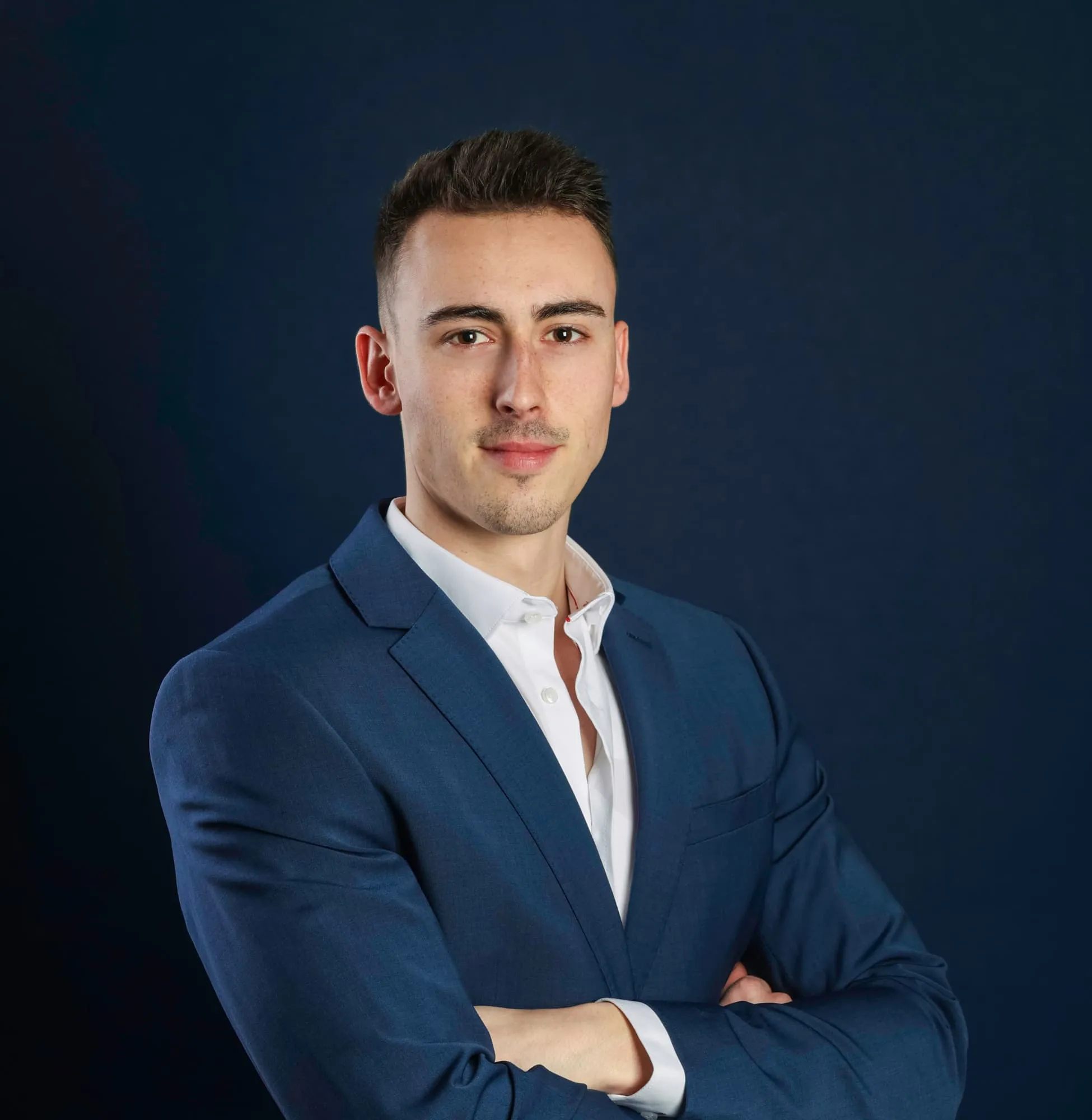Young man with short dark hair wearing a blue suit jacket and white shirt, standing with arms crossed against a dark blue background.