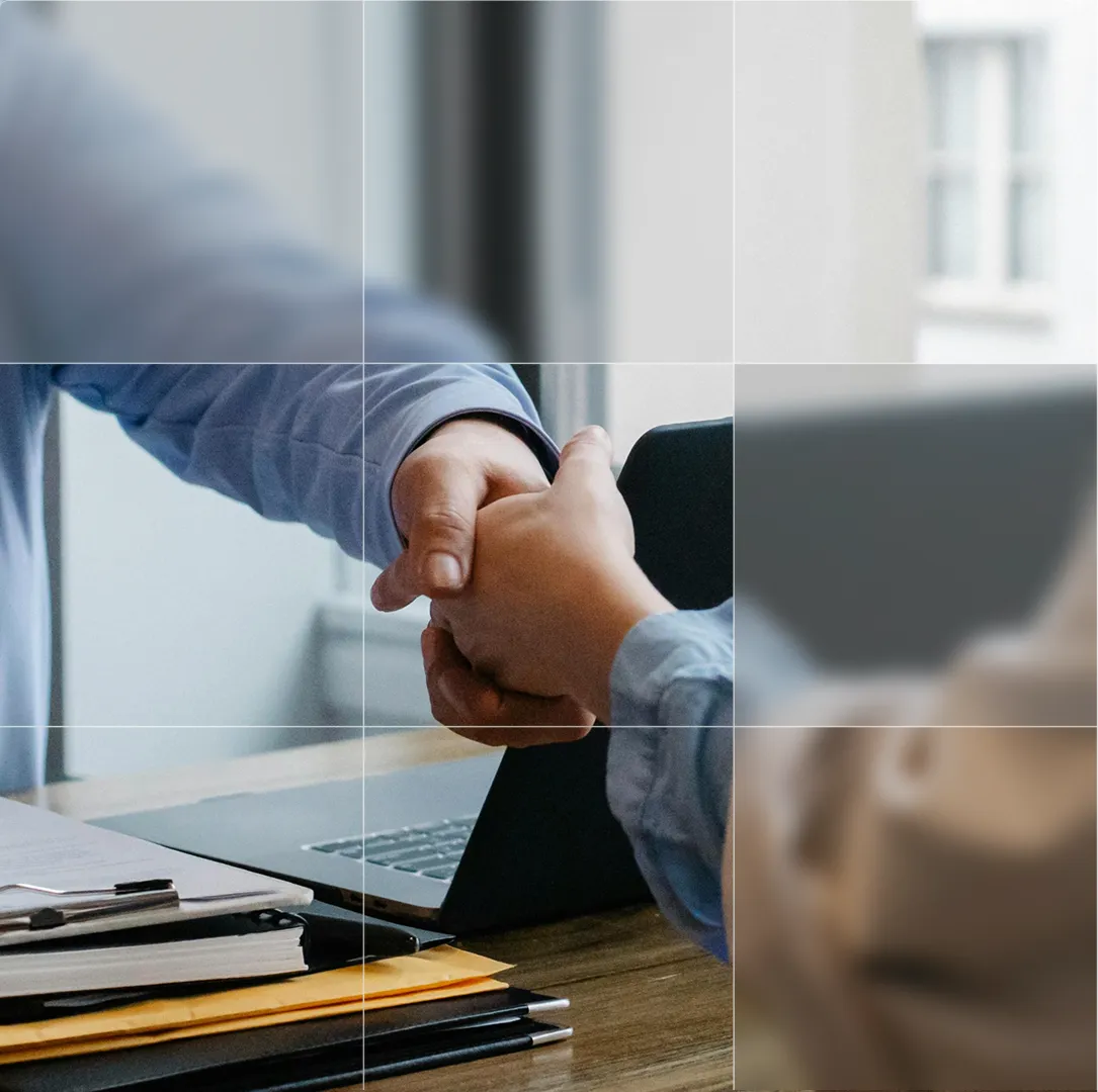 Two people shaking hands across a desk with a laptop and stacked folders.