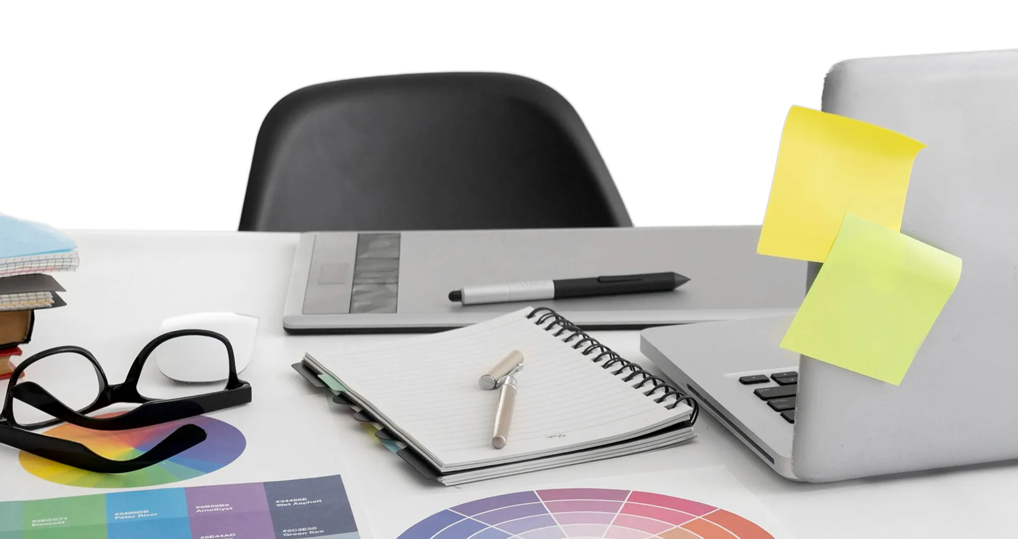 Desk with a laptop, sticky notes, glasses, color swatches, a notebook with a pen on top, and a drawing tablet with a stylus.