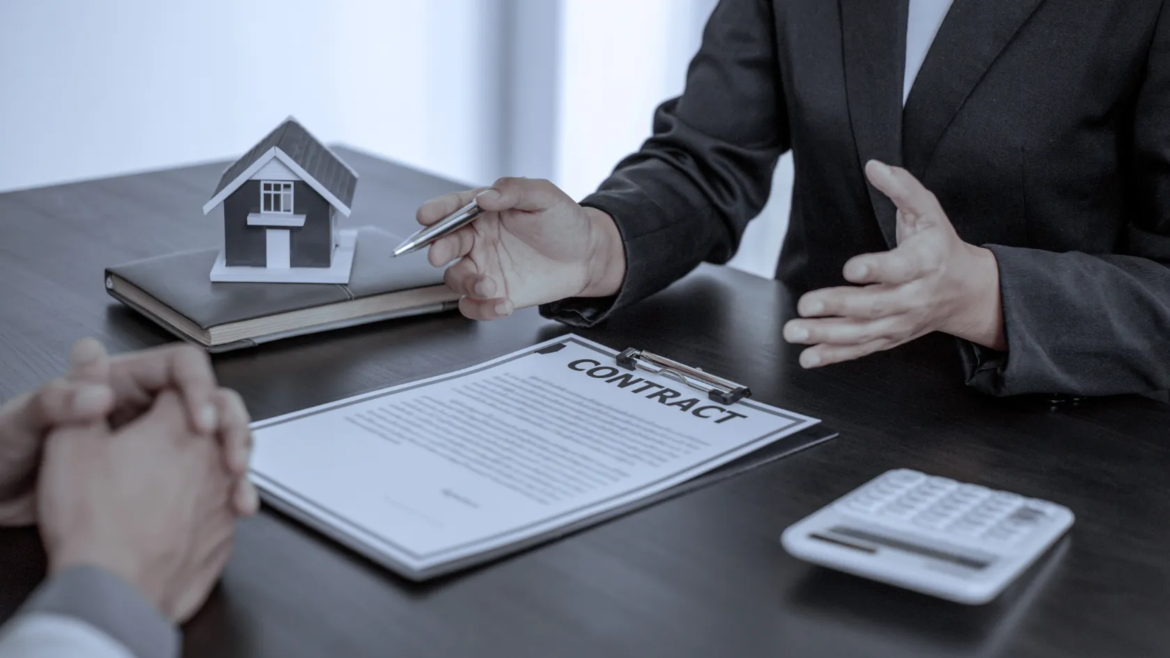 Two people discussing a contract on a desk with a small house model and a calculator nearby.
