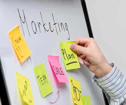 Hand placing a sticky note labeled 'Plan' on a whiteboard with other colorful sticky notes and the word 'Marketing' written above.
