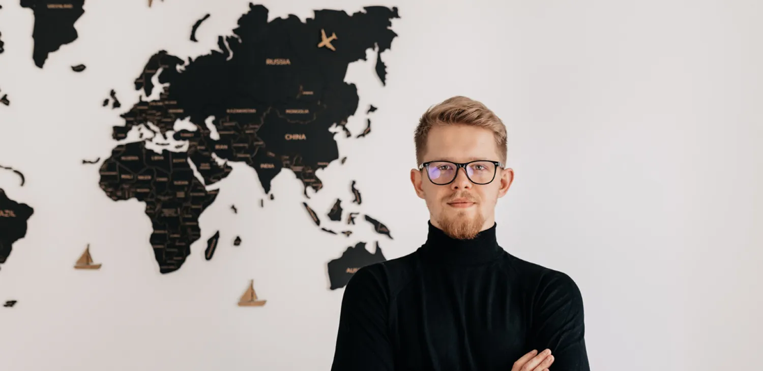 Man with glasses and beard wearing a black turtleneck standing with arms crossed in front of a wall-mounted world map.