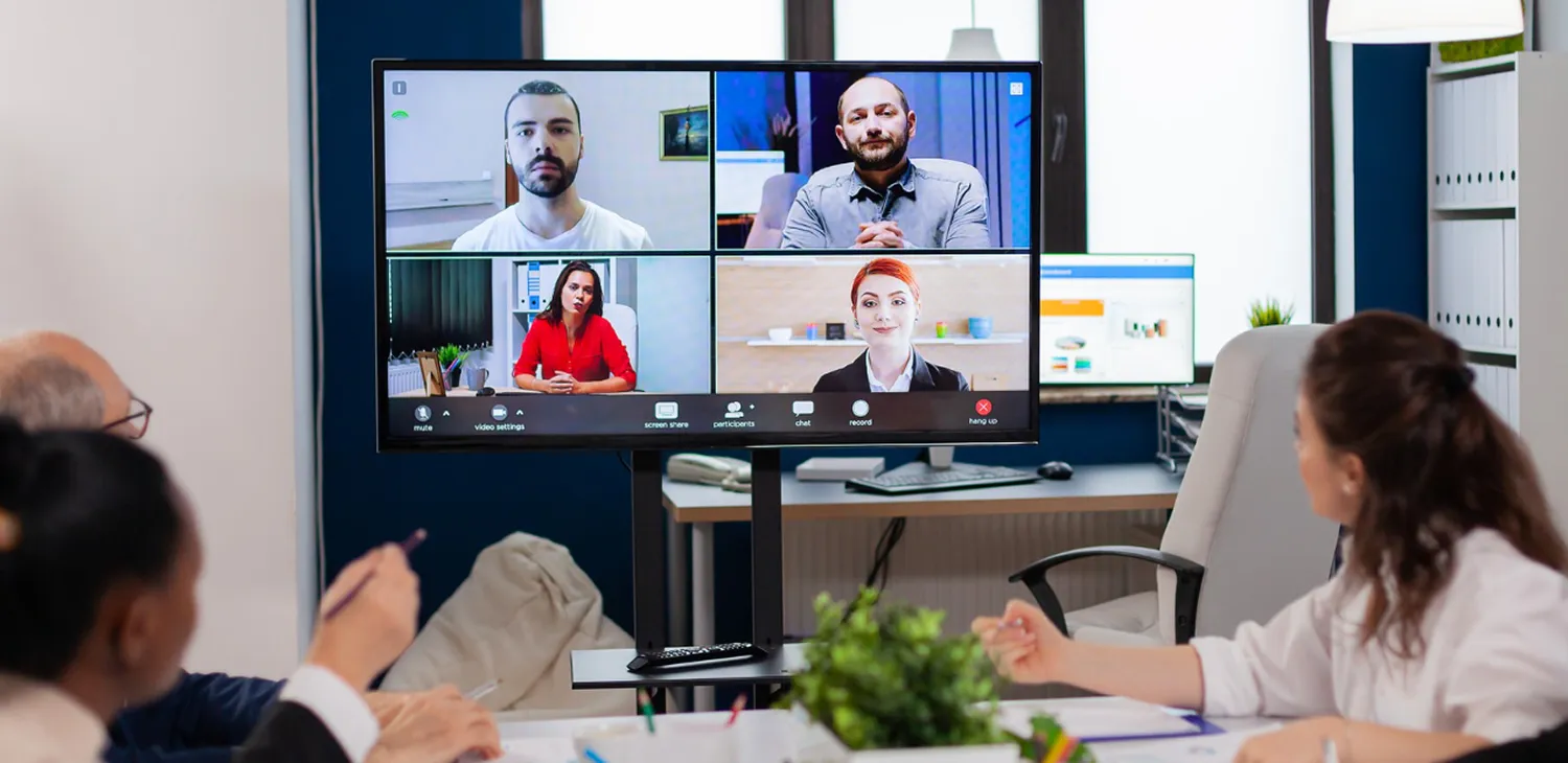 Four colleagues engaged in a video conference call displayed on a large monitor in an office setting.