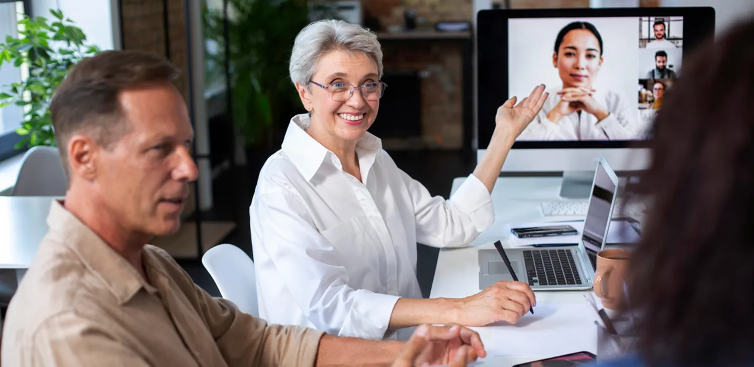 Smiling woman in white shirt pointing toward a computer screen showing a video conference with four people.