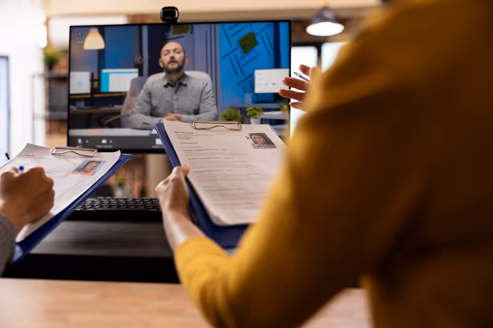 Two interviewers holding resumes on clipboards while conducting a virtual job interview with a man on a computer screen.