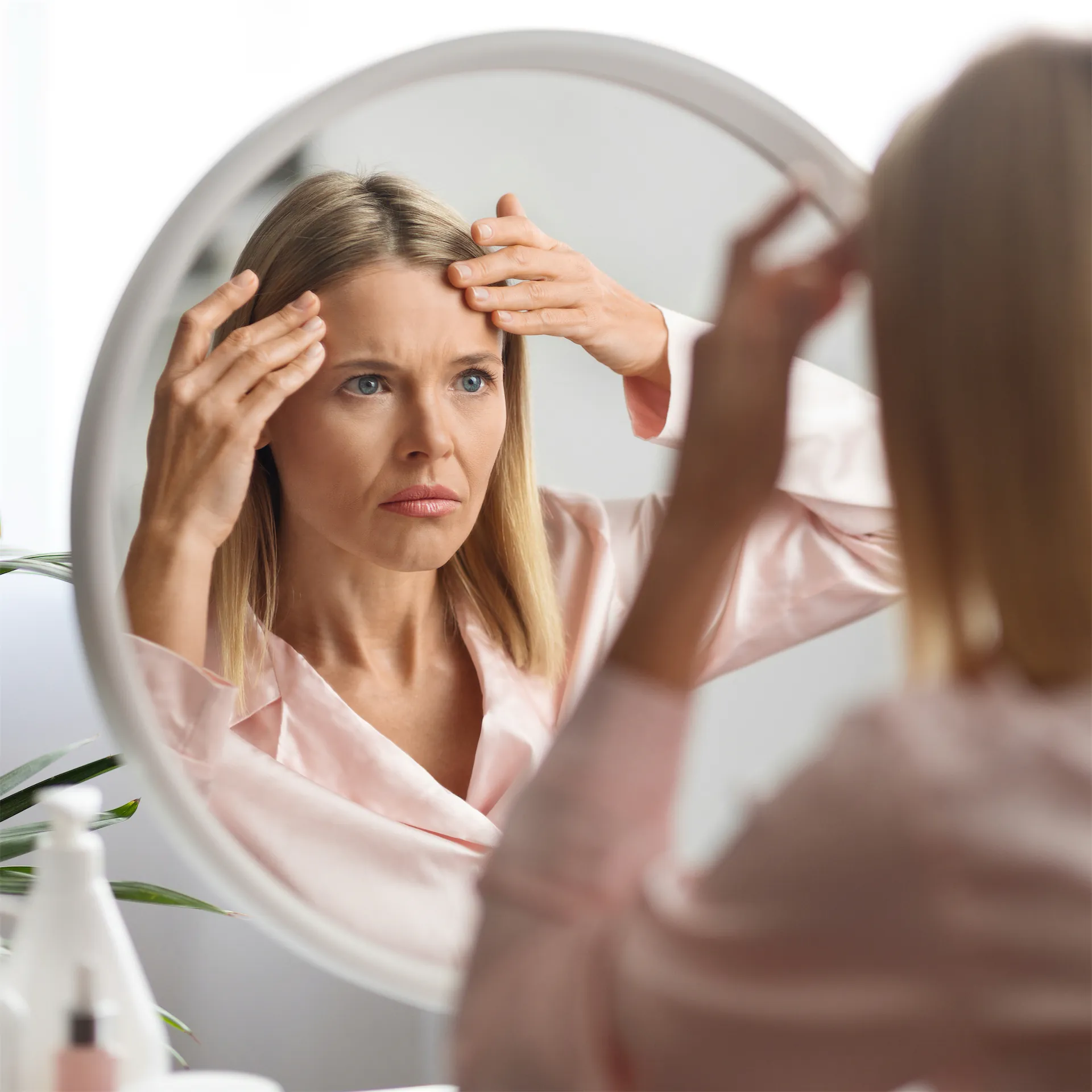 A middle-aged woman frowning at the fine lines and wrinkles she sees on her forehead in the reflection of her bathroom mirror