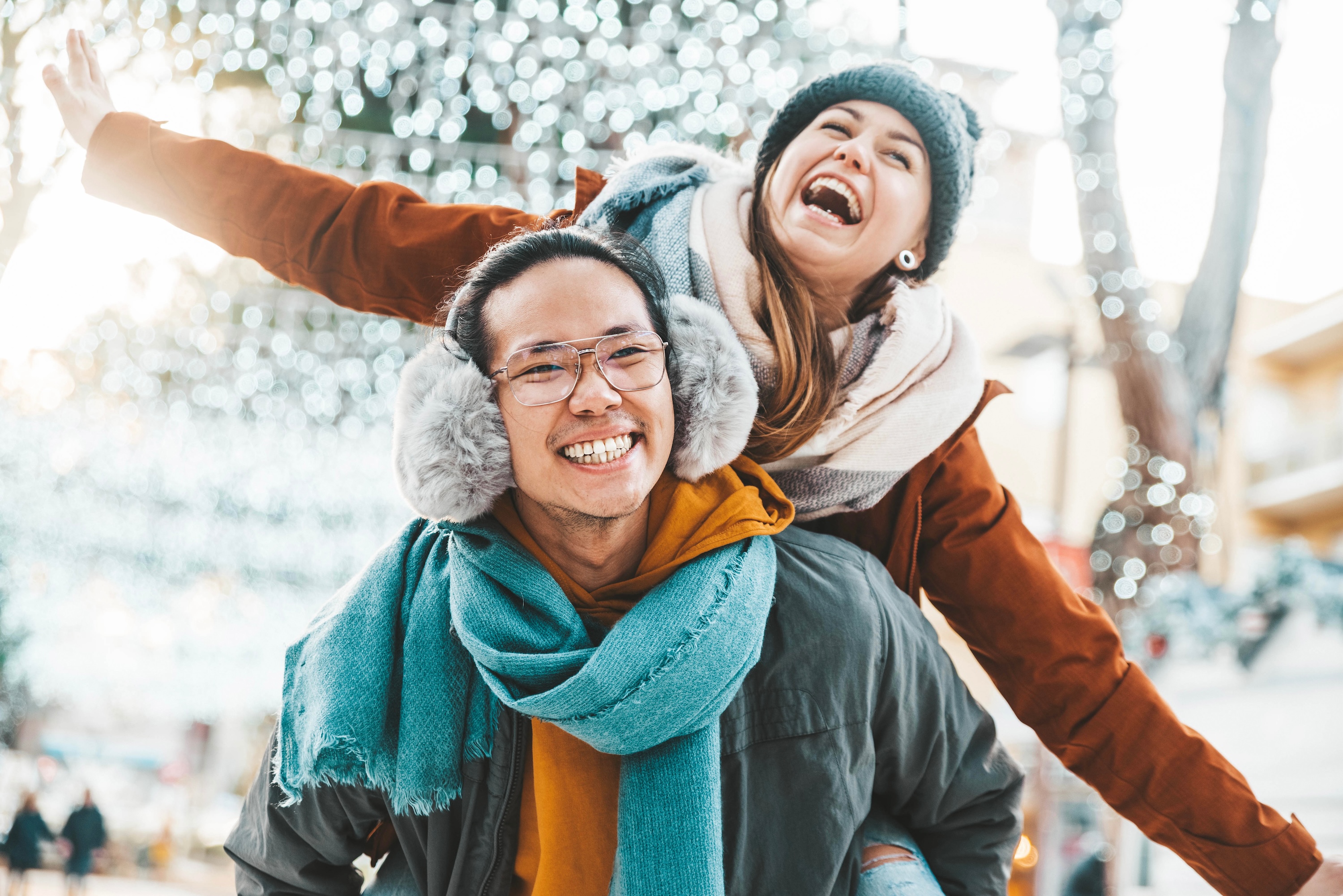 A happy man giving a piggyback ride to a laughing woman in winter gear against a backdrop of festive holiday lights, representing the cheerful community atmosphere at The Grand at Riverside in Evans, CO.