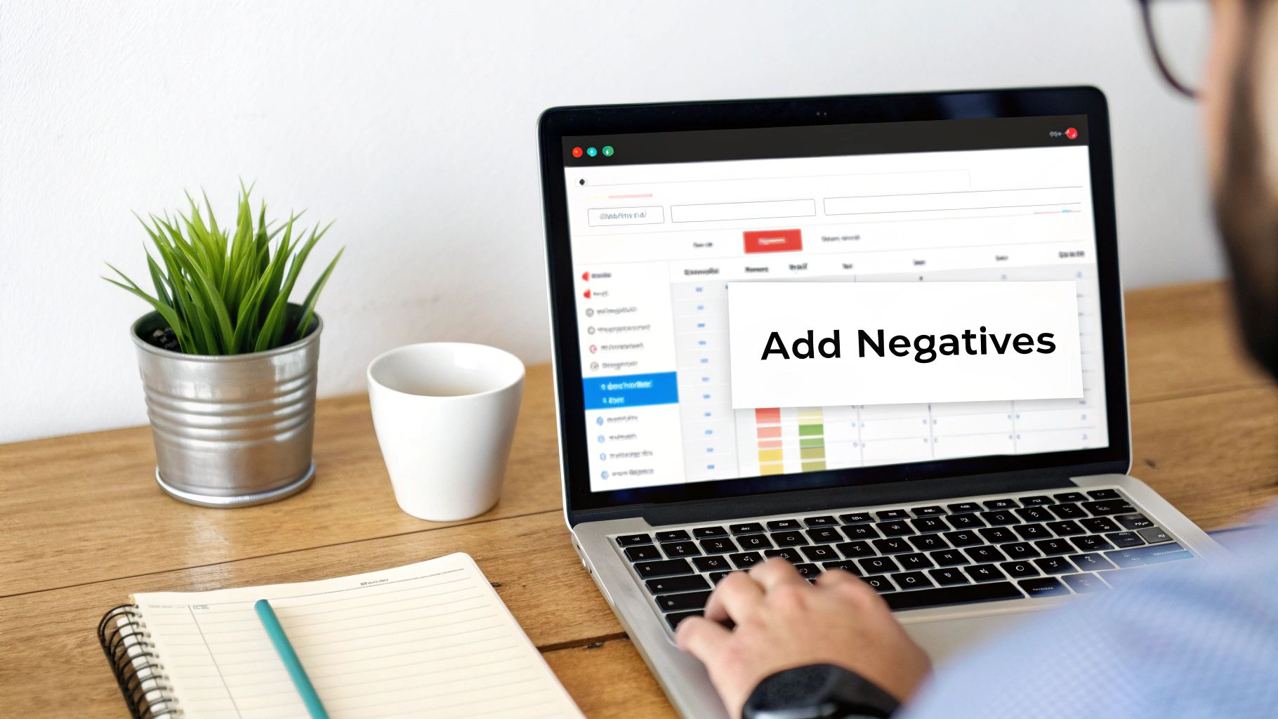 A person typing on a laptop, adding negative keywords to a digital campaign, with office items on a desk.