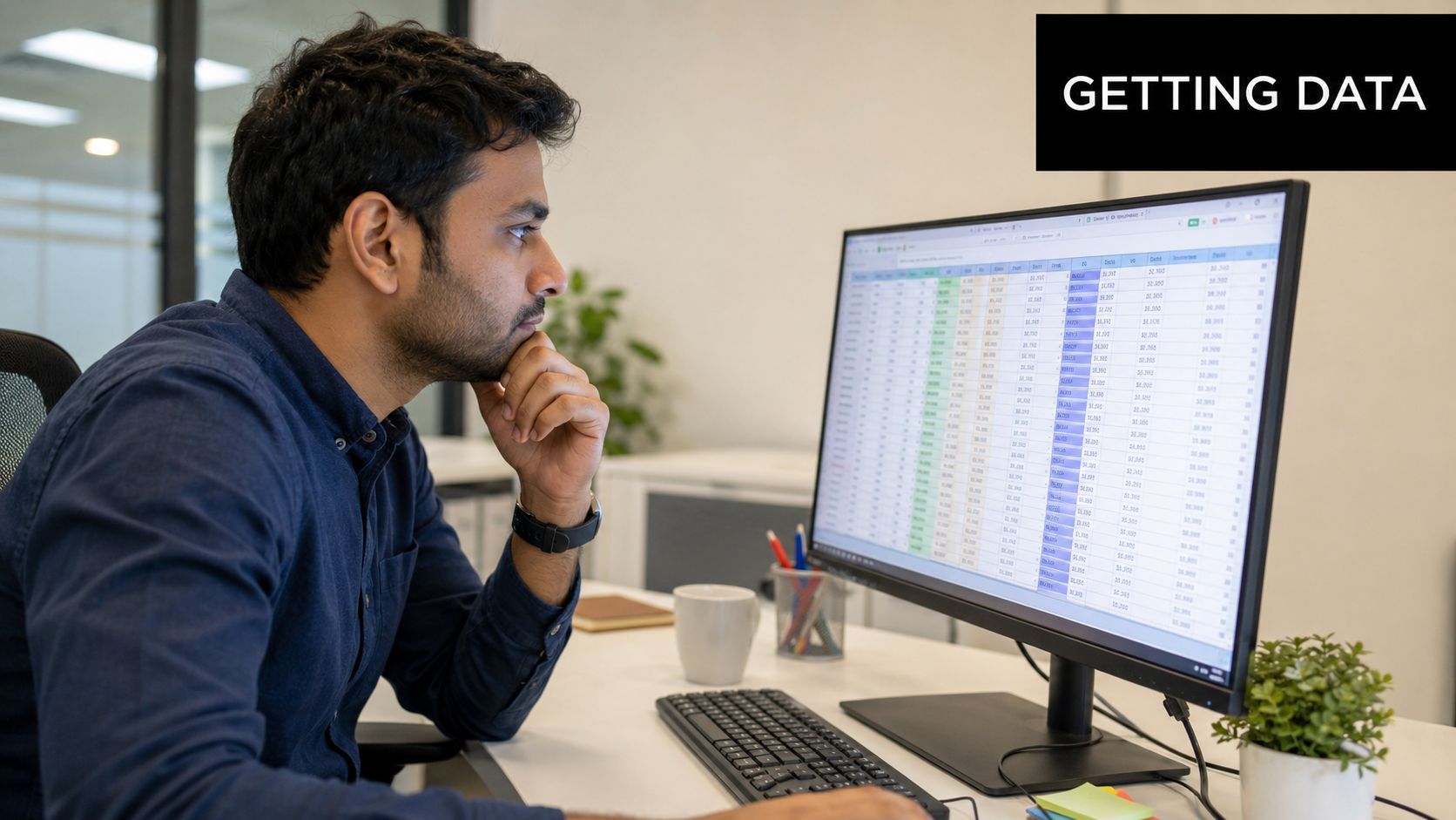 A professional man sitting at a desk looking intently at a computer screen displaying complex spreadsheet data.