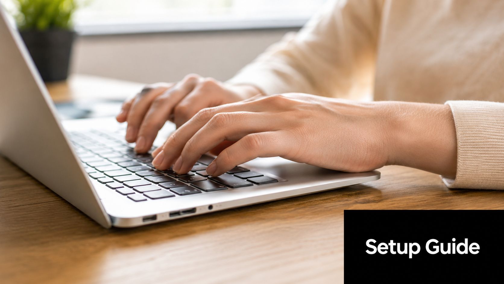 A person wearing a beige sweater typing on a silver laptop on a wooden desk.