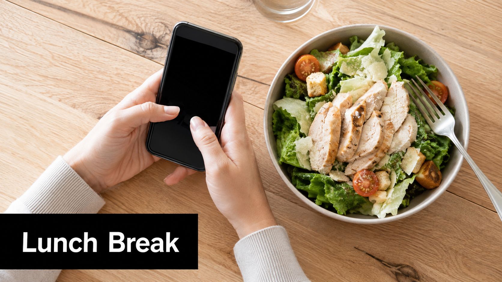 A person sitting at a wooden table holding a smartphone while eating a fresh chicken salad.