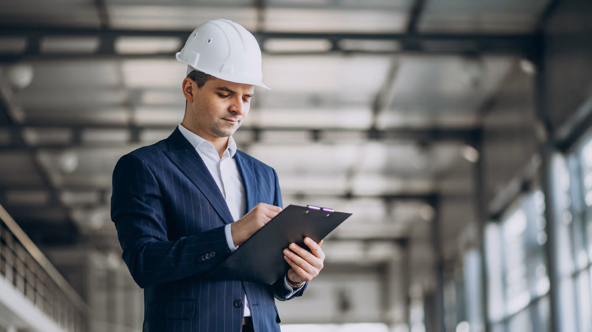 Man wearing a suit and hardhat filling out paperwork on a clipboard in an industrial building.