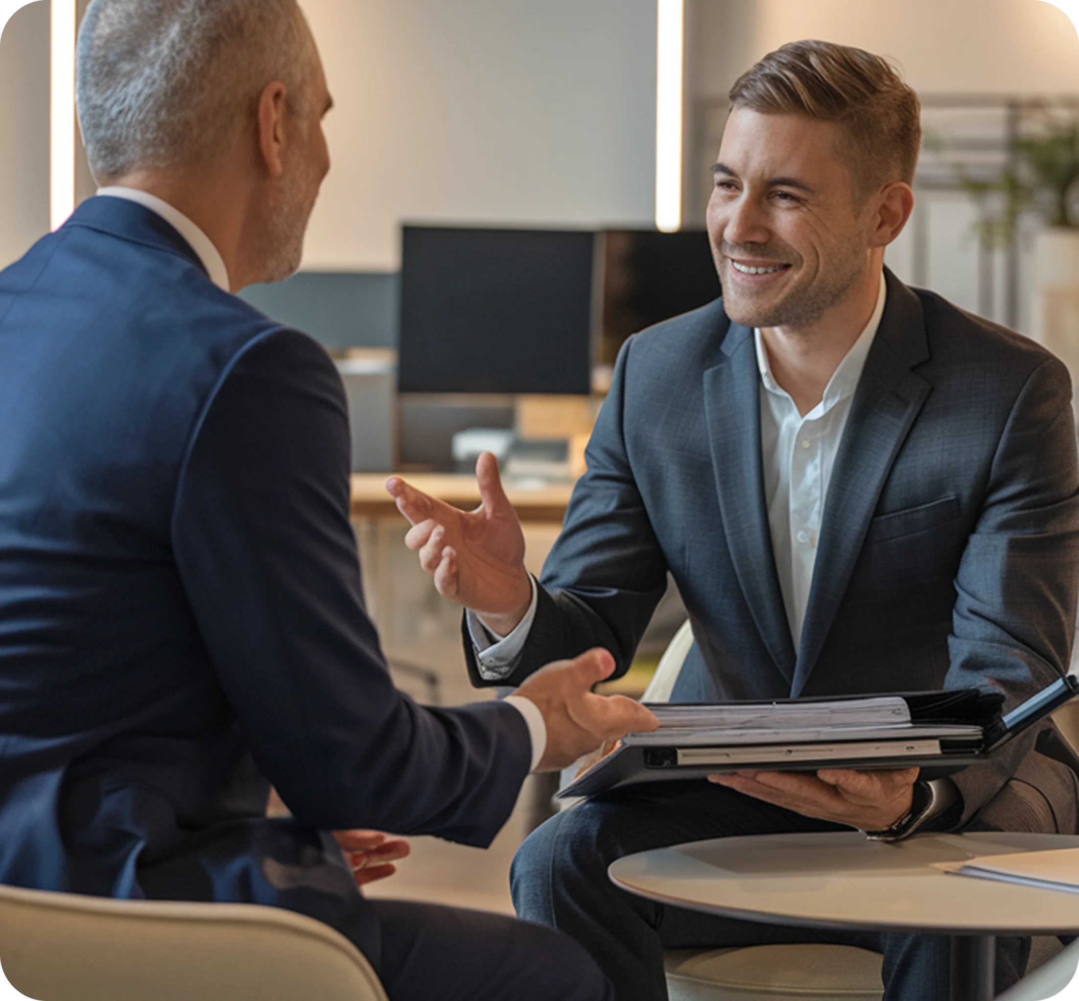 Two men in suits in a meeting, one smiling and holding a folder